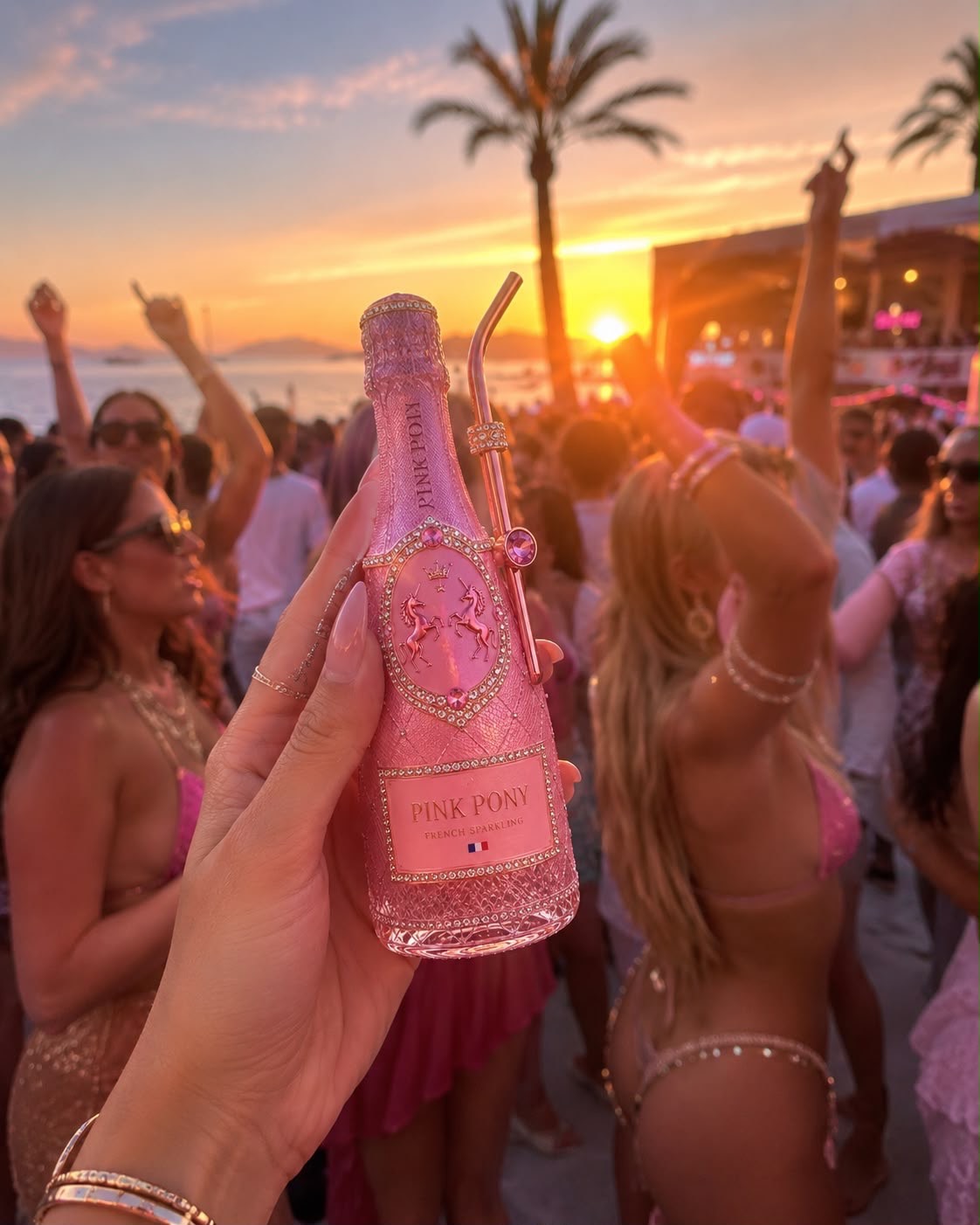 Hand holding a Pink Pony bottle toward the golden sunset sky, crowd dancing below